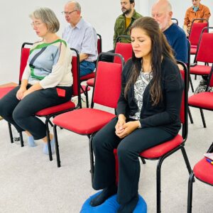 A mixed-age group meditating on chairs in a bright hall, a woman sitting on a blue cushion in the foreground with her eyes closed