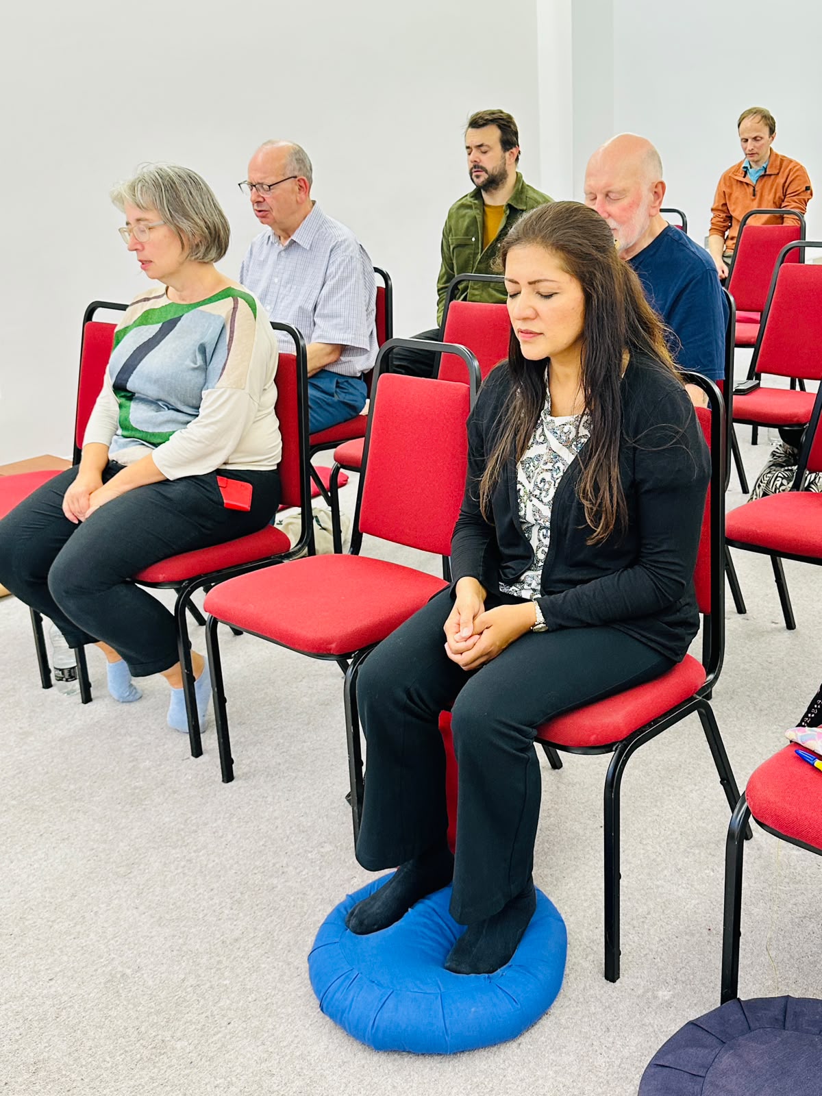 A mixed-age group meditating on chairs in a bright hall, a woman sitting on a blue cushion in the foreground with her eyes closed