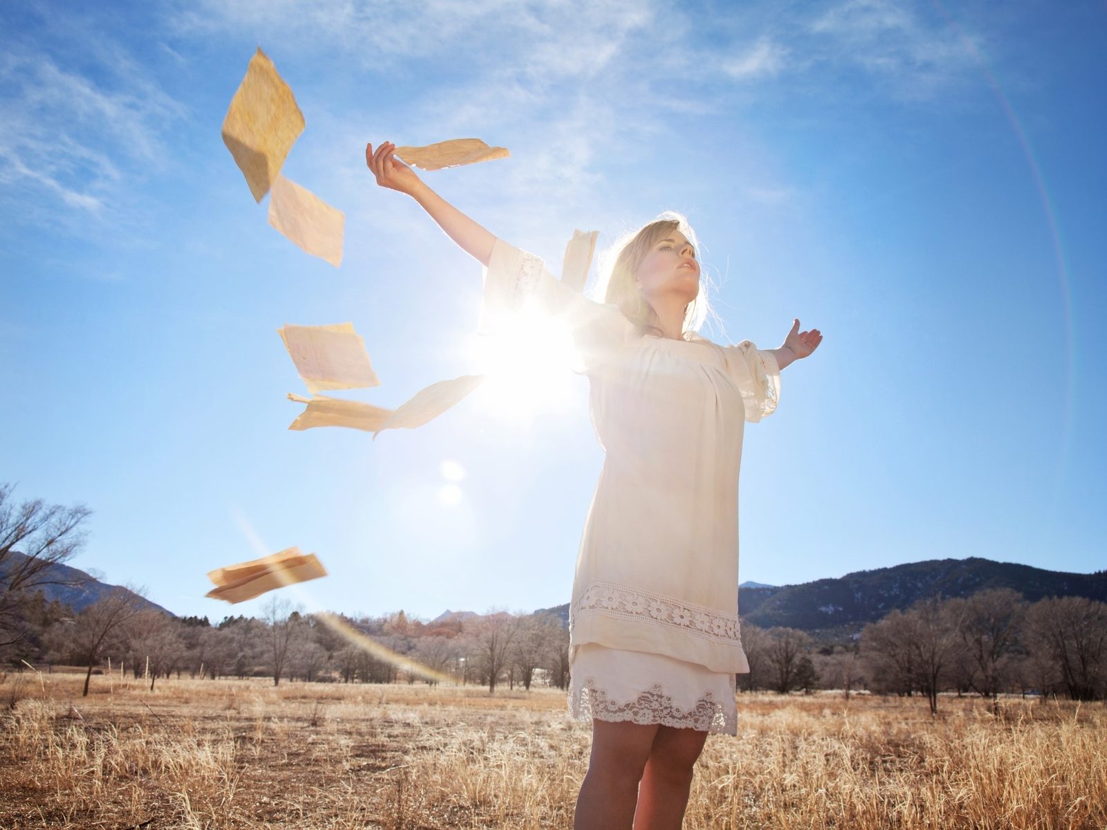 Woman in a white dress standing in a sunlit winter field, arms outstretched, releasing sheets of paper into a bright blue sky