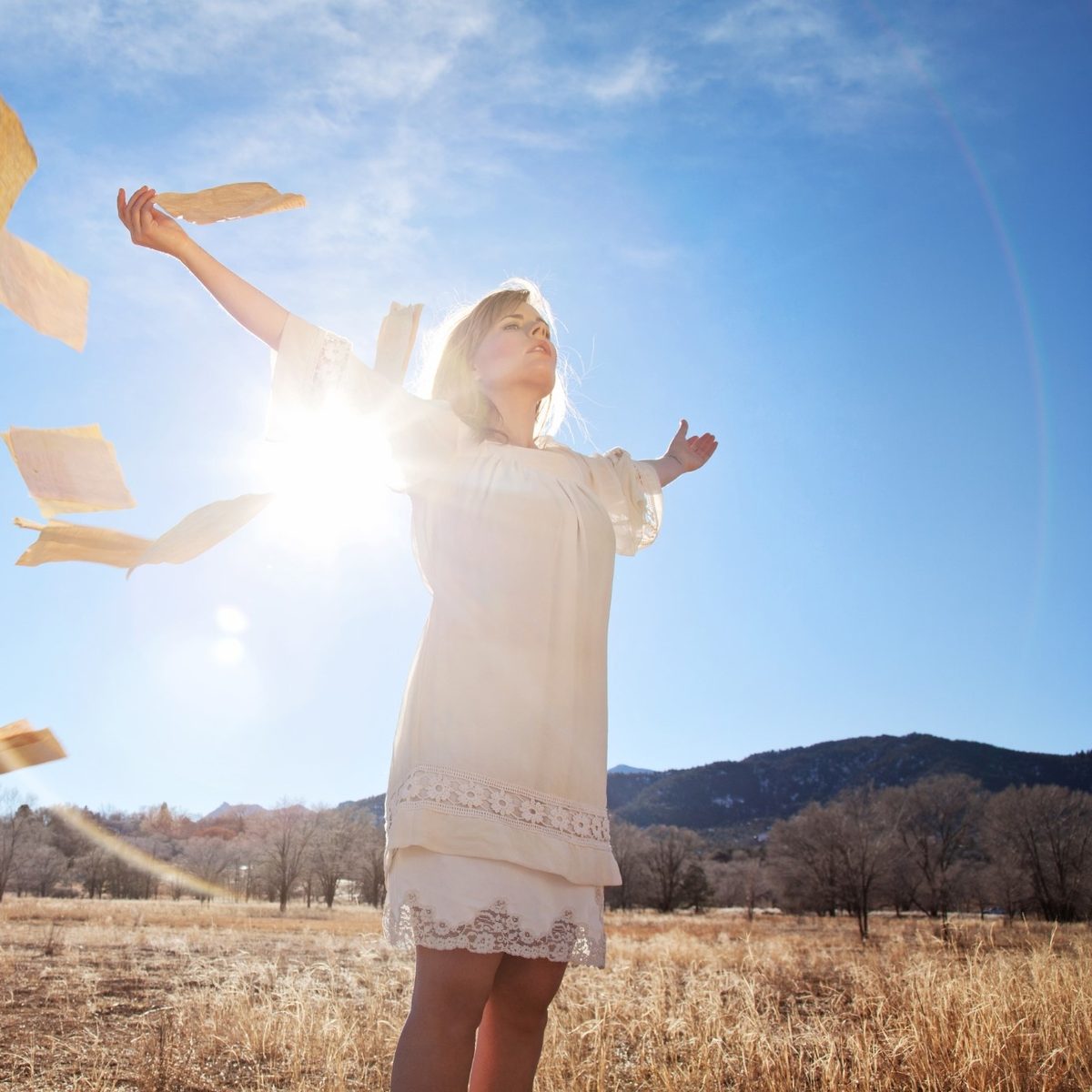 2026-11-07-letting-go-of-hurt-square Woman in a white dress standing in a sunlit winter field, arms outstretched, releasing sheets of paper into a bright blue sky