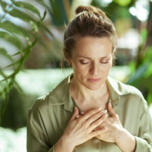 Woman with her eyes closed and her hands resting on her chest, surrounded by green plants