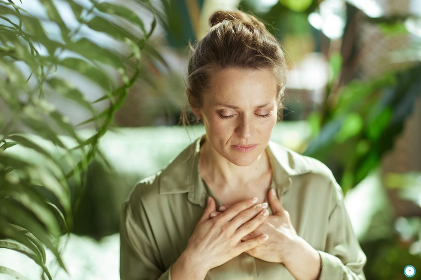 2026-11-21-being-kind-to-yourself Woman with her eyes closed and her hands resting on her chest, surrounded by green plants