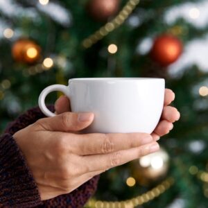 Hands holding a white cup in front of a decorated Christmas tree with warm lights and red and gold baubles.