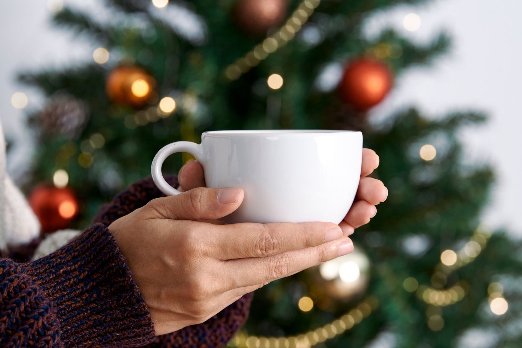 2026-12-05-calm-in-the-christmas-rush Hands holding a white cup in front of a decorated Christmas tree with warm lights and red and gold baubles.
