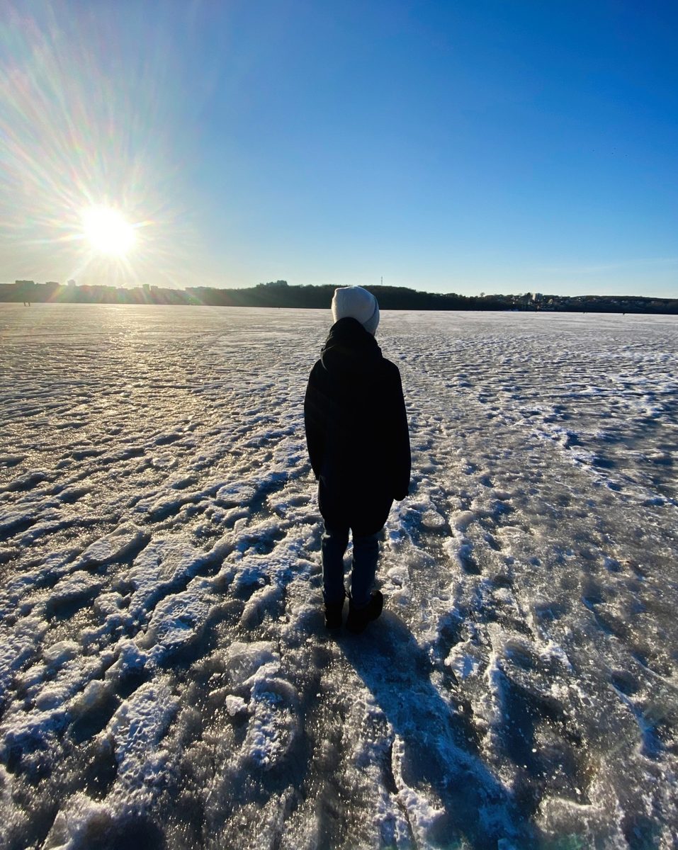 Person in a woolly hat standing on a frozen lake, looking towards the low winter sun